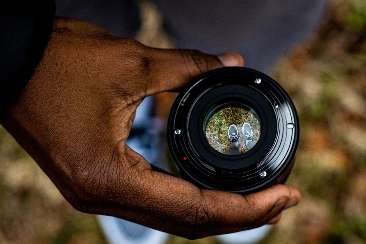 A close-up shot of a hand holding a camera lens with sneakers in view outdoors, creating a unique perspective.