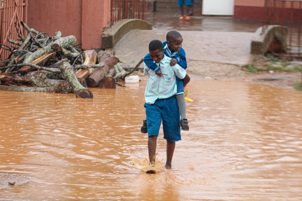 Two children wade through a flooded street, with one giving the other a piggyback ride.