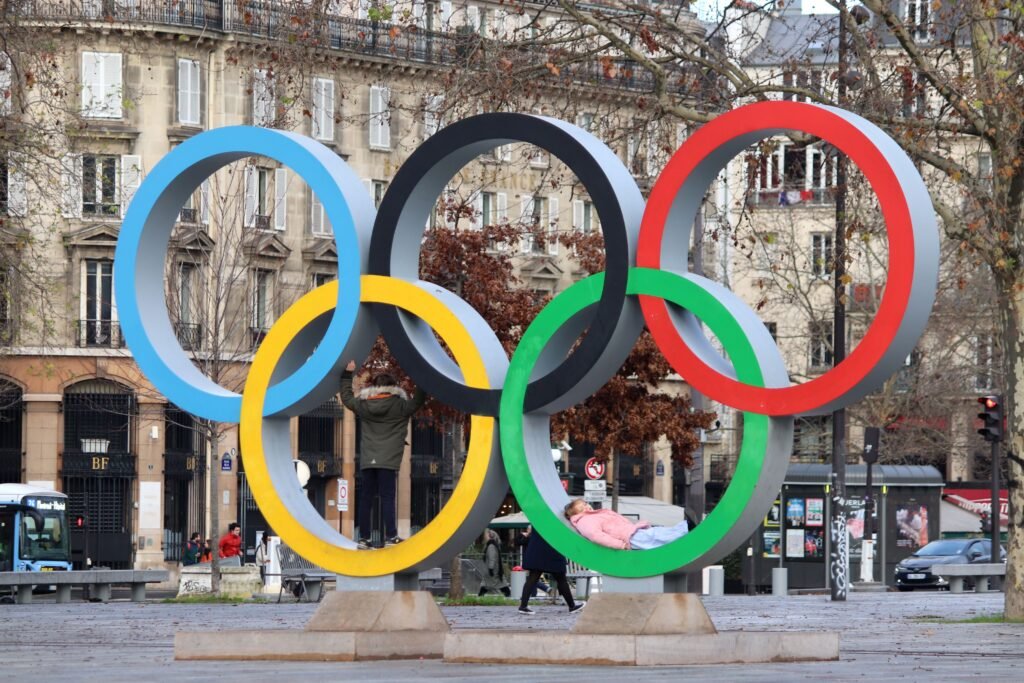 Olympic Rings statue with Paris architecture backdrop, symbolizing sport unity.